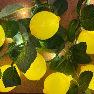 Può includere: Una ghirlanda decorativa di luci a forma di limone giallo con foglie verdi. Le luci sono disposte su una superficie marrone, creando un'esposizione luminosa e allegra. Perfetta per aggiungere un tocco d'estate.