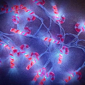 Puede incluir: Una cadena de luces navideñas con forma de bastones de caramelo. Las luces son rojas y blancas con un diseño en espiral, e iluminan con un brillo suave y cálido. El conjunto está sobre un fondo oscuro.