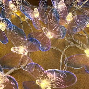 Pode incluir: Uma série de luzes decorativas com formas de borboletas translúcidas em tons de azul e rosa. Cada borboleta tem uma pequena luz LED branca quente. As luzes são enfiadas em um fio transparente, criando uma exibição caprichosa.