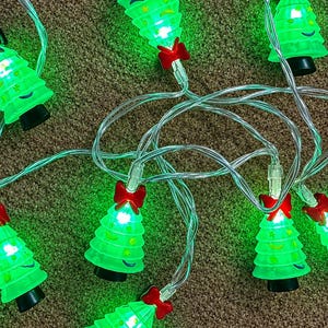 May include: A string of LED lights featuring translucent green Christmas tree-shaped covers. Each tree is accented with a small red bow at the top and decorated with yellow star and moon shapes. The lights are connected by a clear electrical cord.