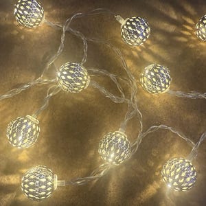 Può includere: Stringa luminosa decorativa con globi argentati traforati. Ogni globo emette una luce bianca calda. Le luci sono montate su un filo trasparente, perfette per la decoro interno o esterno. Ideale per creare un'atmosfera festosa.