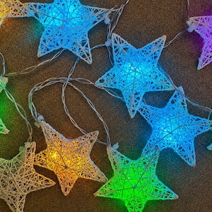 May include: A string of star-shaped fairy lights with a woven design. The stars are illuminated in a variety of colours, including blue, green, yellow, and purple. The lights are strung on a thin, silver wire, and are arranged on a brown surface.