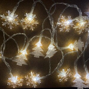 Puede incluir: Una cadena de luces navideñas con bombillas transparentes en forma de copo de nieve y árbol de Navidad. Las luces están ensartadas en un cable transparente y emiten un brillo blanco cálido. Decoración festiva.