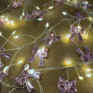 Op de afbeelding: Een decoratieve lichtslinger met kleine, doorschijnende roze strikversieringen. De lampjes zitten op een dunne, zilverkleurige draad en de strikken zijn gelijkmatig verdeeld. De achtergrond is een getextureerd, bruin oppervlak en de lampjes zijn verlicht.