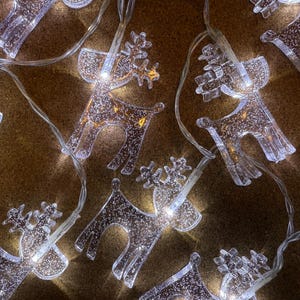 May include: A string of clear, glittery reindeer-shaped Christmas lights. The lights are illuminated, casting a warm glow. The reindeer have detailed antlers and are connected by a thin, silver wire. The background is a textured, brown surface.