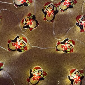 May include: A string of Christmas lights featuring penguin-shaped ornaments. Each penguin is black and white with a red Santa hat, scarf, and a gift. The lights are on a thin, clear wire.