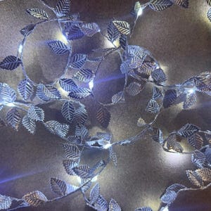May include: A silver leaf garland with small white LED lights. The leaves have a detailed veining pattern and are strung together with a thin wire. The lights are evenly spaced along the garland, creating a soft glow.