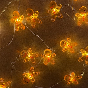 May include: A string of warm, glowing gingerbread man-shaped Christmas lights. The lights are a translucent orange color, with small red accents. The string is thin and silver, and the background is a textured brown.