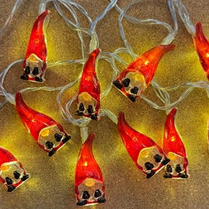 Op de afbeelding: Een decoratieve lichtslinger met kleine kaboutervormige lampen. Elke kabouter heeft een rode hoed, een geel gezicht en een zwarte baard en laarzen. De lampjes zijn geregen aan een transparante draad en geven een warme gloed.