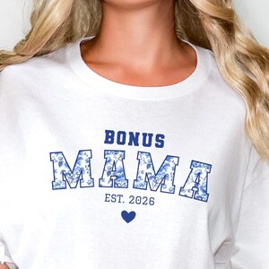 Könnte beinhalten: Ein weißes Kurzarm-T-Shirt mit dem blauen Schriftzug BONUS MAMA, dessen Buchstaben mit einem Blumenmuster gefüllt sind, sowie dem Text EST. 2026 und einem kleinen blauen Herzsymbol unter dem Schriftzug. Das Shirt wird mit einer hellblauen Jeans kombiniert.