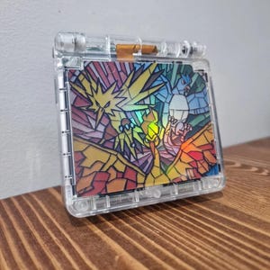 May include: A clear, transparent Game Boy Advance SP shell featuring a stained-glass design with the legendary Pokémon Zapdos, Articuno, and Moltres. The artwork is vibrant with yellow, red, blue, and green hues. The handheld device is displayed on a wooden surface.