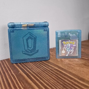 Op de afbeelding: Een blauwe glittercase en een spelcartridge. De case heeft een geometrisch ontwerp. De spelcartridge heeft een Pokémon-label met de tekst "Crystal Version". De items staan op een houten oppervlak.