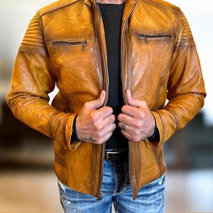 Könnte beinhalten: Eine gealterte, goldbraune Lederjacke mit Reißverschluss und mehreren Taschen. Die Jacke hat gesteppte Schulterdetails und einen klassischen Biker-Stil. Das Model trägt eine blaue Jeans und ein schwarzes T-Shirt.