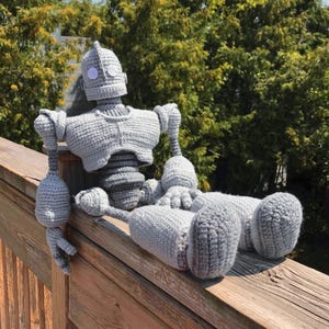 以下が含まれることがあります： 丸い頭、2つの大きな目の円、詳細な体を持つ、灰色の編み物ロボット人形。 人形は木の柵に座っています。 人形の高さは約30cmです。