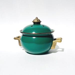 May include: A round, deep green enameled pot with a matching lid featuring a small brass-colored knob. The pot has two brass-colored side handles and a dark rim. The lid includes small ventilation holes near the top center of the dome.