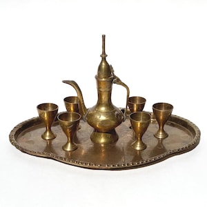Puede incluir: Un juego de té de latón vintage, que incluye una tetera decorativa con un pico largo y seis vasos pequeños, dispuestos sobre una bandeja ovalada. El juego tiene un diseño ornamentado y está sobre un fondo blanco.