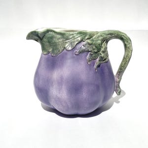May include: A ceramic pitcher in the shape of an eggplant. The pitcher is a muted purple color with a green handle and rim, detailed with leaf-like designs. The pitcher is on a white background.