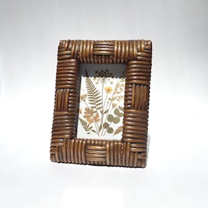 May include: A rectangular picture frame constructed from dark brown woven rattan reeds. The frame features a repeating pattern of horizontal and vertical reed segments. Inside the frame is a print of pressed botanical specimens including ferns.