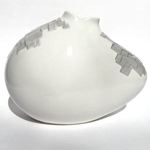 Puede incluir: Un jarrón de cerámica blanco con una forma redondeada única y un patrón gris texturizado. El jarrón tiene un acabado brillante y un diseño asimétrico distintivo. El patrón gris se asemeja a una serie de pequeños bloques rectangulares.