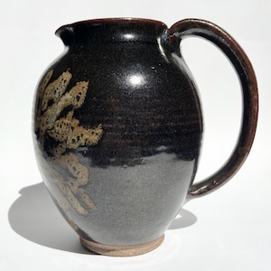 Puede incluir: Una jarra de cerámica marrón oscuro con un cuerpo redondeado y un asa grande. La jarra tiene un acabado brillante y un diseño decorativo abstracto en tonos dorados. La base es de un color más claro, sin esmaltar.