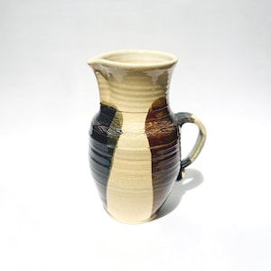 May include: A ceramic pitcher featuring a cream-colored base with vertical stripes in dark brown and black. The vessel has a rounded body, a flared rim with a pouring spout, and a curved handle on the side. The surface shows horizontal ridges.