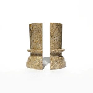 Puede incluir: Un par de sujetalibros cil&iacute;ndricos hechos de piedra f&oacute;sil en tonos beige y bronceado. Cada pieza presenta una superficie lisa y pulida con inclusiones org&aacute;nicas visibles y un dise&ntilde;o de base escalonada y ensanchada para mayor estabilidad.