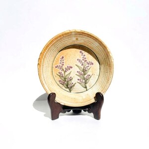 May include: A round ceramic plate with a speckled beige and tan glaze, featuring a hand-painted design of two sprigs of purple lavender flowers in the center. The plate has a ridged rim and is displayed on a dark brown wooden stand.