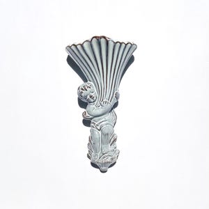 May include: A wall-mounted ceramic vase featuring a sculpted figure of a cherub holding a large, fluted shell-shaped vessel. The piece is finished in a glossy, pale blue-grey glaze with dark brown accents highlighting the molded details.