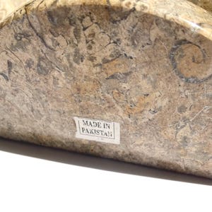 Puede incluir: Un sujetalibros semicircular hecho de piedra fosil&iacute;fera pulida con un patr&oacute;n moteado en tonos bronceado, marr&oacute;n y gris con inclusiones org&aacute;nicas visibles. Una peque&ntilde;a etiqueta rectangular blanca en el frente muestra el texto MADE IN PAKISTAN.