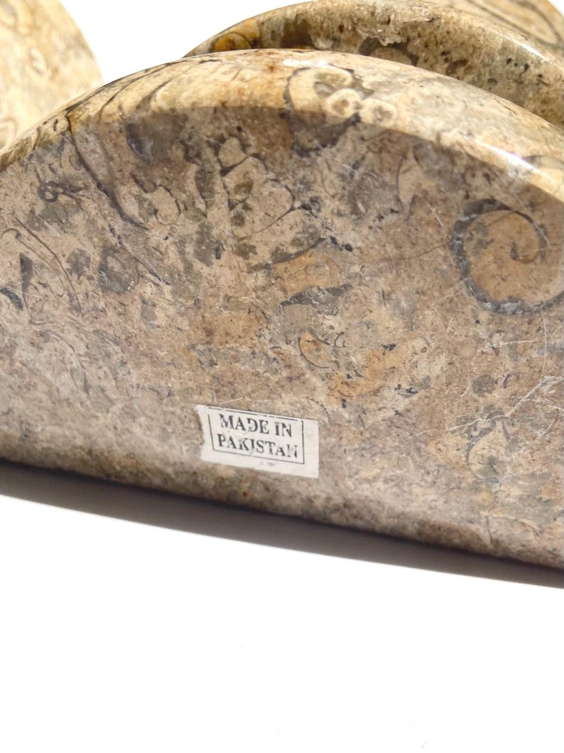 Puede incluir: Un sujetalibros semicircular hecho de piedra fosil&iacute;fera pulida con un patr&oacute;n moteado en tonos bronceado, marr&oacute;n y gris con inclusiones org&aacute;nicas visibles. Una peque&ntilde;a etiqueta rectangular blanca en el frente muestra el texto MADE IN PAKISTAN.