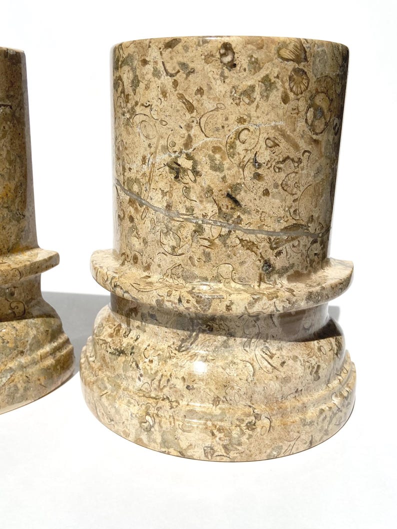 Puede incluir: Dos pedestales cil&iacute;ndricos de piedra con un patr&oacute;n fosil&iacute;fero en tonos beige y bronceado. Cada pieza tiene una superficie lisa y pulida con inclusiones org&aacute;nicas visibles y un dise&ntilde;o de base redondeada y escalonada. La piedra muestra vetas naturales.