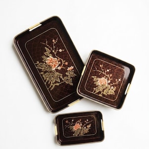 May include: A set of three dark brown rectangular and square serving trays with rounded corners and light-colored woven handles. Each tray features a gold-toned floral design with pink blossoms and a fine, crackled grid pattern across the surface.