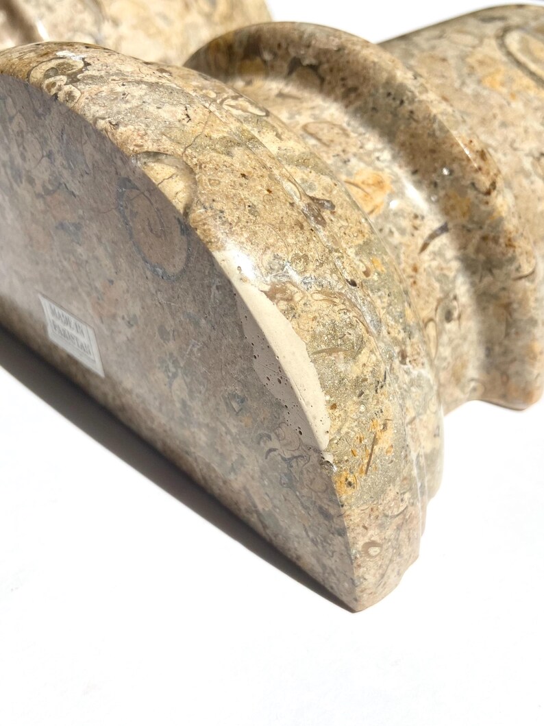 Puede incluir: Un sujetalibros semicircular pulido hecho de piedra fosil&iacute;fera en tonos beige y bronceado. La superficie presenta un patr&oacute;n natural complejo de formas org&aacute;nicas incrustadas e inclusiones minerales. Una etiqueta blanca indica Made in Pakistan.
