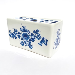 May include: A rectangular white ceramic flower brick vase featuring blue floral patterns on the front and side. The top surface contains multiple circular openings for arranging individual flower stems in a structured display.