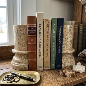 Puede incluir: Dos sujetalibros cil&iacute;ndricos de piedra beige con textura moteada flanquean una fila de cinco libros de tapa dura titulados The Silmarillion, Classical Architecture, A History of Stone Carving, Curated Collections y Antiques and Curiosities.