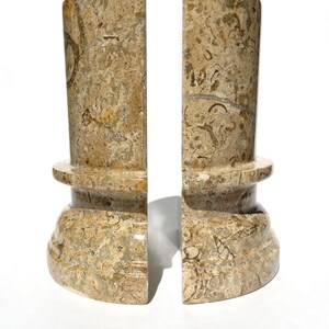 Puede incluir: Un par de sujetalibros cil&iacute;ndricos hechos de piedra caliza fosil&iacute;fera pulida en tonos beige y bronceado. Cada pieza presenta una base ancha y redondeada con un reborde horizontal, mostrando un patr&oacute;n natural de fragmentos de conchas incrustados.