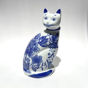 May include: A white ceramic cat figurine decorated with blue floral patterns. The cat is in a seated position with its tail wrapped around its base. It features blue painted ears, eyes, whiskers, and a blue collar around its neck.