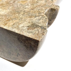 Puede incluir: Un objeto de piedra pulida con una superficie moteada en tonos beige, marr&oacute;n y gris que presenta una textura densa y fosilizada. La pieza tiene un borde grueso y redondeado y una muesca distintiva en el lateral con un patr&oacute;n natural.
