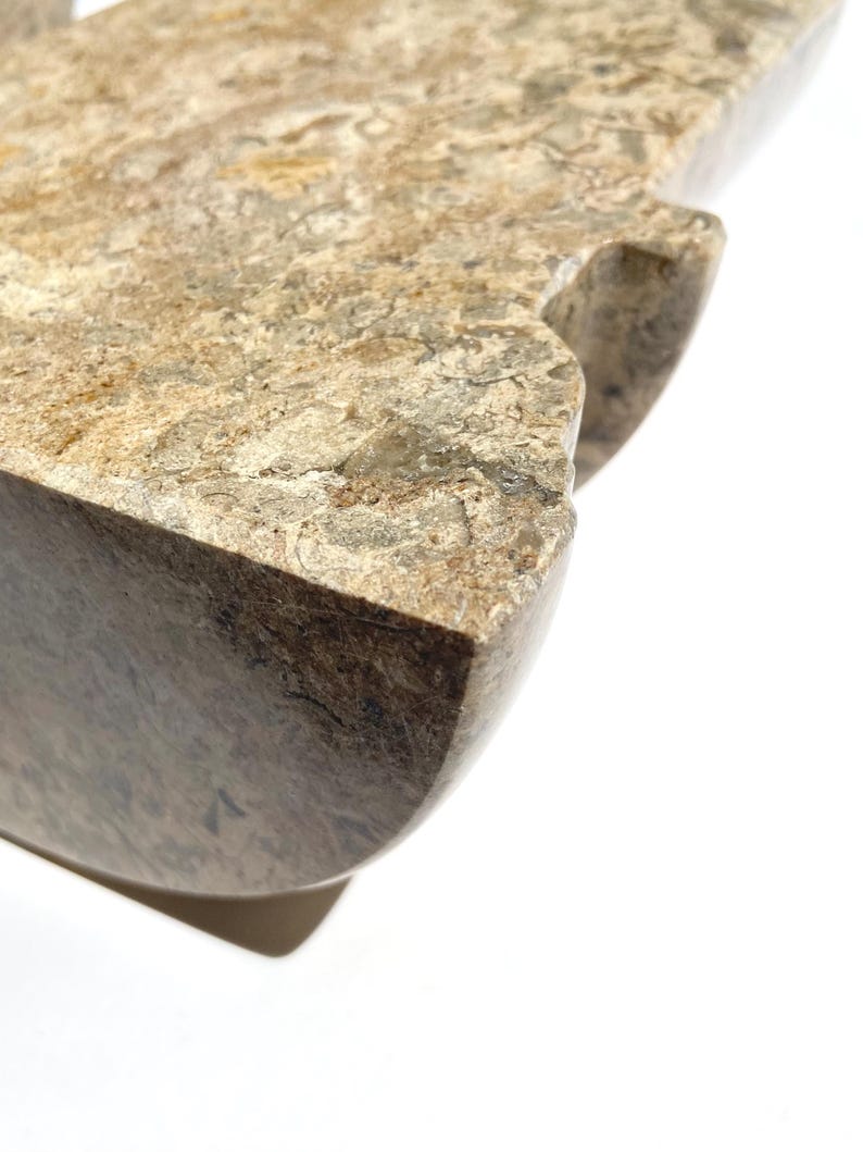 Puede incluir: Un objeto de piedra pulida con una superficie moteada en tonos beige, marr&oacute;n y gris que presenta una textura densa y fosilizada. La pieza tiene un borde grueso y redondeado y una muesca distintiva en el lateral con un patr&oacute;n natural.