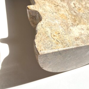 Puede incluir: Un objeto de piedra pulida con una superficie moteada en tonos beige y bronceado que presenta inclusiones f&oacute;siles visibles. La pieza tiene un borde curvo y una muesca distintiva en el lateral, mostrando una textura densa y tonos naturales.