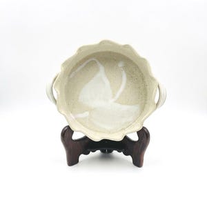 May include: A round, off-white ceramic bowl with a scalloped rim and two side handles. The interior features a speckled texture and a white, abstract, brushstroke-style design. The bowl is displayed on a dark brown, three-legged wooden stand.