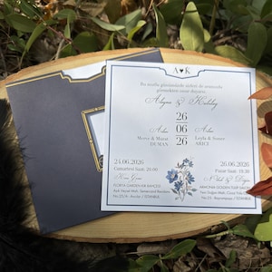 May include: A wedding invitation with a navy blue envelope and a white card. The card features the names of the couple, the date, and floral accents. The invitation is set on a wooden surface with greenery.