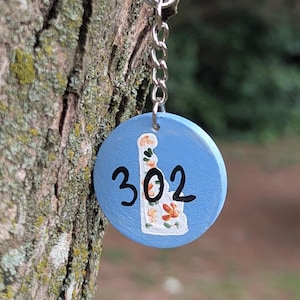 May include: A round, blue keychain with the number "302" and a floral design. The keychain is attached to a silver keyring and chain. The background is a tree trunk.