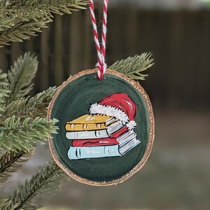 May include: A wooden Christmas tree decoration featuring a painted design of a stack of books and a Santa hat. The round ornament has a green background and a red and white string. It is hanging on a Christmas tree.
