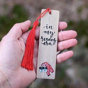May include: Wooden bookmark with a red tassel. The bookmark features the text "in my reading era" in black script. A small illustration of books and a scarf is at the bottom. The bookmark is held in a hand.