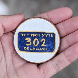 May include: A round wooden ornament featuring a painted design of a Delaware license plate. The plate is blue with yellow and white accents, and the text reads "THE FIRST STATE 302 DELAWARE". The ornament is held in a hand.
