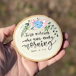 May include: A round wooden slice with painted floral accents in pink, blue, and green. The text "His mercies are new every morning" is written in black script, with the reference "LAM. 3:23".
