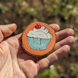 May include: A hand-painted wooden slice featuring a cupcake design. The cupcake has a blue base, white frosting, and a red cherry on top. The wood slice has an orange border and a natural wood grain.