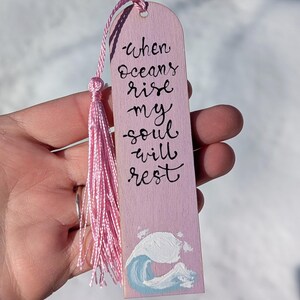 May include: A light pink wooden bookmark featuring the black hand-lettered text When oceans rise my soul will rest. The bookmark includes a light pink tassel attached to the top and a painted blue and white ocean wave at the bottom.