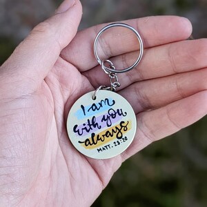 May include: A circular keyring featuring the handwritten phrase "I am with you always" in black, with pastel stripes of blue, yellow, and purple. The text "MATT. 28:20" is below. The keyring is attached to a silver ring.